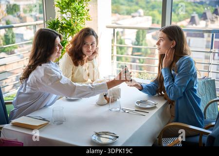 Eine junge Frau, die im Restaurant einen Handy-Bildschirm für ihre Freundinnen zeigt Stockfoto