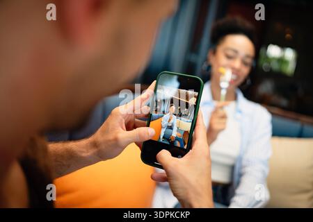 Ein vielseitiges Paar mit Feinschmecker genießt ein zwangloses Abendessen und einen Telefonmoment, bei dem sonnige Vibes und Drinks geteilt werden Stockfoto