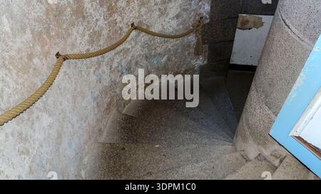 Eine schmale Wendeltreppe aus Stein mit einem Seilgeländer zieht sich gegen eine verwitterte Mauer nach oben und erinnert an alten architektonischen Charakter und rustikalen Charme. Stockfoto