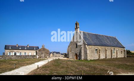 Eine Dorfkirche aus Stein mit einem hohen Turm steht unter einem klaren blauen Himmel, umgeben von traditionellen Häusern in einer ruhigen ländlichen Umgebung. Stockfoto