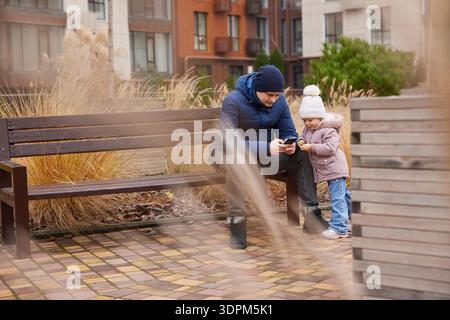 Vater und Tochter interagieren mit Mobiltelefonen, während sie auf einer Bank in einem Stadtpark sitzen Stockfoto