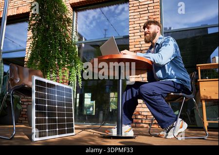 Mann arbeitet am Laptop an einem runden Tisch im Freien, mit einem Glas Eiskaffee. Solarpanel in der Nähe, der nachhaltigen, modernen, umweltfreundlichen Arbeitsbereich betont, der Technologie und Nachhaltigkeit vereint. Stockfoto