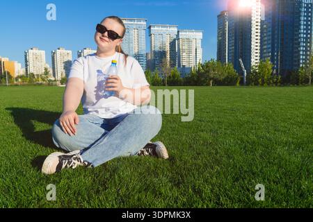 Junge blonde Frau in weißem T-Shirt, die auf grünem Rasen sitzt und Wasserflasche mit modernen Wohnhochhäusern und blauem Himmel auf dem Hintergrund hält Stockfoto