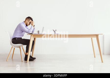 Mann, der allein an einem Schreibtisch sitzt, mit einem Laptop, der Zeichen von Stress in einem hellen Raum zeigt Stockfoto