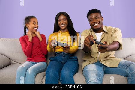 Die Familie genießt gemeinsam die Game Night auf einer Couch mit den Game Controllern in den Händen und einem Lächeln auf den Gesichtern Stockfoto