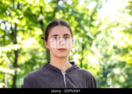 Eine junge Frau porträtiert draußen in einem sonnendurchfluteten Park mit einem grauen Hoodie mit neutralem Ausdruck Stockfoto
