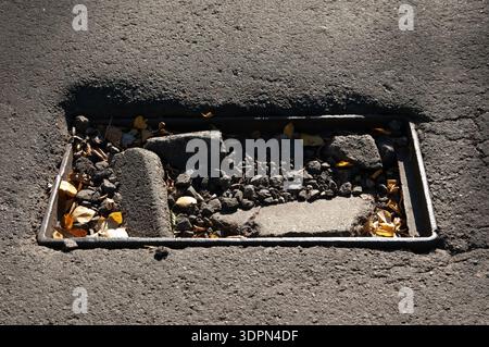 Alte verlassene Sturmrinne in Asphalt unter hellem Sonnenlicht. Der Abfluss ist mit Asphaltbrocken, Kies und herabfallenden Blättern verstopft. Kein Gitter, nur ein Metall fr Stockfoto