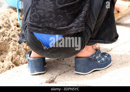 Offene Nahaufnahme einer Person, die sich auf einem Steinweg hockt, mit markanten blauen Jeanssandalen und schwarzer Kleidung. Urban Lifestyle Fotografie Stockfoto