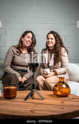 Zwei lächelnde Frauen erstellen Online-Inhalte mit Smartphone und Lavalier-Mikrofonen und sitzen während eines Live-Streams auf einem Sofa Stockfoto