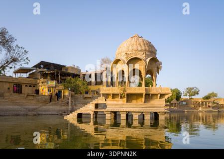 Jaisalmer, Indien - 28. März 2022: Ein kunstvoller Sandsteinpavillon mit Kuppeldach steht auf Säulen im Wasser am Gadisar Lake, mit Stufen, Gebäuden, Bäumen und Menschen entlang des Seeufers im Hintergrund. Stockfoto