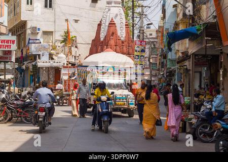 Udaipur, Indien - 23. März 2022: Ein dekorierter Lastwagen fährt eine enge Straße mit Fußgängern, Motorrädern, Motorrollern, Geschäften und sichtbaren Hindi-Schildern entlang und fährt tagsüber vor einem Hindutempel mit einem rot-weißen Shichara. Stockfoto