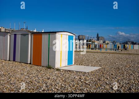 Le Havre, Frankreich - 25. August 2020: Reihen von kleinen Strandhütten mit vertikalen farbigen Streifen stehen an einem Kieselstrand, wo Menschen laufen und moderne Stadtgebäude im Hintergrund unter einem klaren blauen Himmel sichtbar sind. Stockfoto
