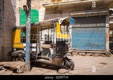 Jodhpur, Indien - 26. März 2022: Eine Auto-Rikscha mit gelber und schwarzer Fassade parkt neben einem Versorgungsmast auf einer sonnendurchfluteten Straße vor einem geschlossenen Geschäft mit blauem Metallladen und alten Steinhäusern. Stockfoto