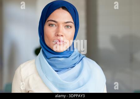 Indische Frau mit marineblauem Hijab und cremefarbenem Pullover, die mit verschwommener Pflanze im Büro nach vorne blickt Stockfoto