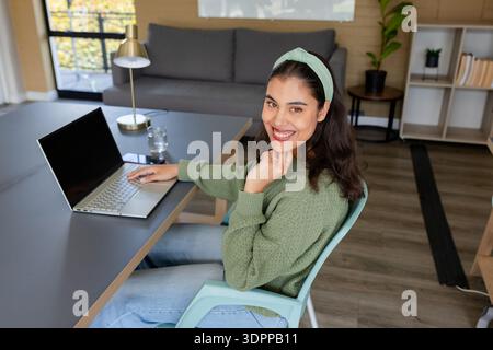 Frau, die am Schreibtisch im Heimbüro sitzt, Laptop und Smartphone am Fenster benutzt, Kopierraum Stockfoto