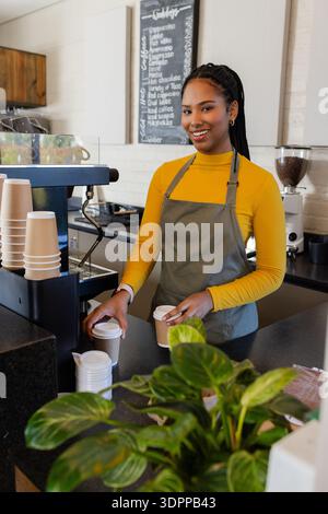 Eine afroamerikanische Barista steht hinter einer Espressomaschine im Café und hält zwei Tassen zum Mitnehmen Stockfoto