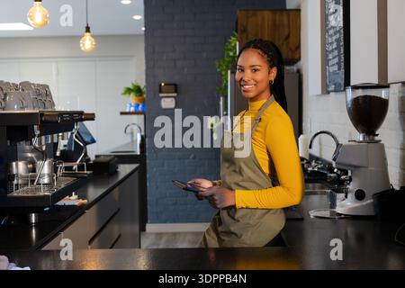 Afroamerikanerin, die eine Schürze trägt und hinter der Theke in einem Café in der Nähe einer Espressomaschine ein Tablet hält Stockfoto