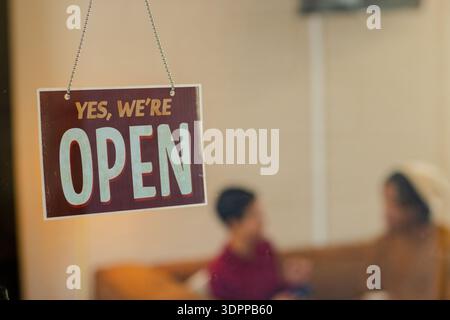 Verschiedene Freunde sitzen auf dem Sofa im Café hinter dem Schaufenster und plaudern in der Nähe eines offenen Schilds und einer Kette Stockfoto