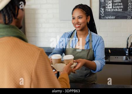Ein vielseitiger Barista mit Schürze und ein afroamerikanischer Mann tauscht zwei Kaffeetassen an der Ladentheke aus Stockfoto