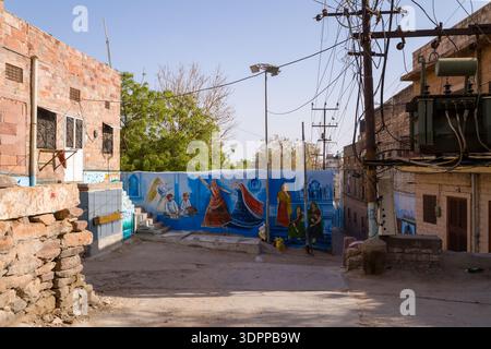 Jodhpur, Indien - 26. März 2022: Eine Straßenszene zeigt ein Wandgemälde auf einer Wand mit traditionellen Rajasthani-Figuren, darunter Musiker und Frauen in bunten Gewändern, mit Wohngebäuden, Versorgungsmasten und Überkopfkabeln. Stockfoto