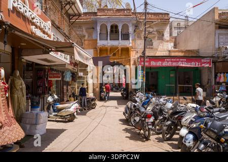 Jodhpur, Indien - 26. März 2022: Zahlreiche Motorräder und Motorroller parken entlang einer sonnendurchfluteten Geschäftsstraße, die von Textilläden, Schaufensterpuppen in traditioneller Kleidung und Schildern in Hindi gesäumt ist, mit einem alten bogenförmigen Tor und historischen Gebäuden im Hintergrund. Stockfoto