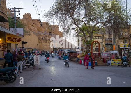 Jaisalmer, Indien - 27. März 2022: Menschen laufen und fahren mit Motorrädern auf einer Straße, die von Geschäften gesäumt ist, darunter einem Bhang-Shop, in der Nähe des Eingangs von Fort de Jaisalmer, mit gelben Sandsteinmauern und Gebäuden im Hintergrund. Stockfoto