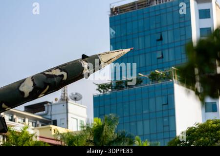 Hanoi, Vietnam - 9. Januar 2020: Nahaufnahme der Nasenpartie einer Rakete mit Tarnfarbe, im Freien im B-52 Victory Museum ausgestellt, mit modernen Gebäuden und Bäumen im Hintergrund. Stockfoto