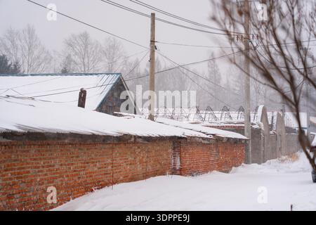 Traditionelle rote Backsteinmauern und Dächer, die während eines Schneesturms in einem ländlichen chinesischen Dorf in Harbin mit starkem Schnee bedeckt sind Stockfoto