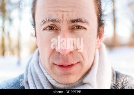 Nahporträt eines Mannes mit Frost an den Augenbrauen und roter Nase in einem Winterpark. Gefrorenes männliches Gesicht, das bei kaltem Wetter draußen in die Kamera schaut Stockfoto