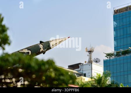 Hanoi, Vietnam - 9. Januar 2020: Nahaufnahme des Nasenabschnitts einer Rakete mit Tarnfarbe, ausgestellt im Freien im B-52 Victory Museum, mit modernen Gebäuden, Bäumen und einem Telekommunikationsturm im Hintergrund. Stockfoto