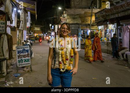 Jodhpur, Indien - 25. März 2022: Eine Frau mit Brille und Rucksack, die mehrere Blumengirlanden trägt, steht lächelnd inmitten einer geschäftigen Nachtstraße mit beleuchteten Geschäften, Hindi-Schildern, Motorrädern und Menschen in traditioneller Kleidung im Hintergrund. Stockfoto
