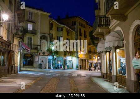 Ville de Verallo, Italien - 27. September 2020: Mehrere Menschen sind nachts auf einer gepflasterten Straße zu sehen, umgeben von leuchtenden gelben und cremefarbenen Gebäuden mit Balkonen, Markisen, Schaufensterfenstern und einem Steindenkmal. Stockfoto