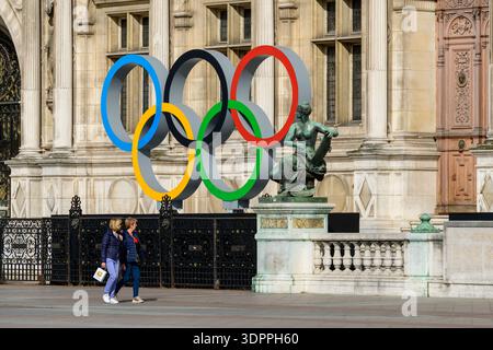 Paris, Frankreich - 20. April 2022: Vor dem Hotel de Ville sind große Skulpturen der Olympischen Ringe und eine sitzende Bronzestatue zu sehen. Zwei Personen gehen an der reich verzierten Fassade des Gebäudes vorbei. Stockfoto