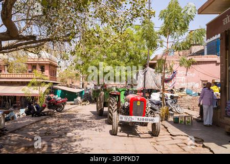 Jodhpur, Indien – 26. März 2022: Ein roter Traktor mit Anhänger und mehreren Motorrädern steht auf einer kopfsteingepflasterten Straße, die von Marktgebäuden, Bäumen und Menschen gesäumt ist, und ein Mann steht in der Nähe eines Ladeneingangs. Stockfoto
