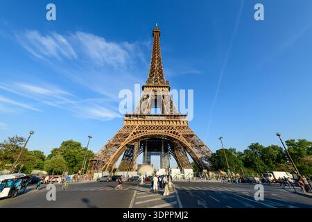 Paris, Frankreich - 10. Mai 2022: Der Eiffelturm ist vom Boden aus zu sehen, mit Menschen zu Fuß, mit dem Fahrrad und mit Fahrzeugen auf der Straße im Vordergrund unter einem klaren blauen Himmel. Stockfoto