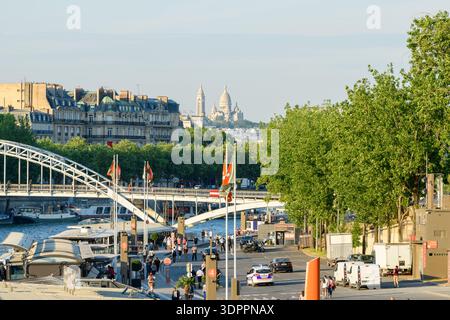 Paris, Frankreich - 10. Mai 2022: Die Fußgängerbrücke Passerelle Debilly überspannt die seine mit Flussbooten, die am Port de la Bourdonnais angedockt sind, die entlang der Uferpromenade laufen und die Basilika Sacré-Cur, die in der Ferne bei klarem Tageslicht sichtbar ist. Stockfoto