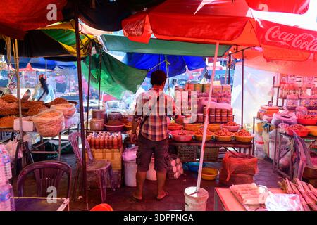 Marche de Kep, Kambodscha - 26. Januar 2020: Ein Verkäufer steht unter bunten Regenschirmen und arrangiert an einem Marktstand im Freien Gläser, Flaschen und Schüsseln mit Lebensmitteln. Um den Stand herum sind Körbe mit getrockneten Waren und verpackten Produkten sichtbar. Stockfoto