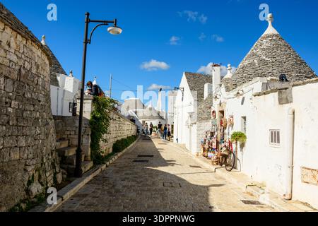 Alberobello, Italien - 17. Oktober 2020: Eine enge Steinstraße, gesäumt von traditionellen weiß getünchten Trullihäusern mit konischen Steindächern, einer Straßenlampe, Topfpflanzen, einem Fahrrad und einer kleinen Straßenverkäuferausstellung unter einem klaren blauen Himmel, bei der mehrere Menschen laufen. Stockfoto