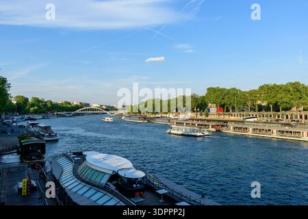 Paris, Frankreich - 10. Mai 2022: Mehrere Flussverbände liegen entlang der seine in Port de la Bourdonnais an, wobei die Fußgängerbrücke Passerelle Debilly im Hintergrund zu sehen ist, von Bäumen gesäumte Böschungen und Menschen, die bei klarem Tageslicht auf der Uferpromenade spazieren gehen. Stockfoto