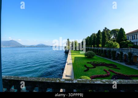 Isola Bella, Italien - 30. September 2020: Das Bild zeigt die formellen Gärten des Palais Borromee mit gepflegten Rasenflächen, Blumenbeeten und dekorativen Blumenbeeten entlang der Uferpromenade, mit Lago Maggiore und fernen Bergen, die unter einem klaren blauen Himmel sichtbar sind. Stockfoto
