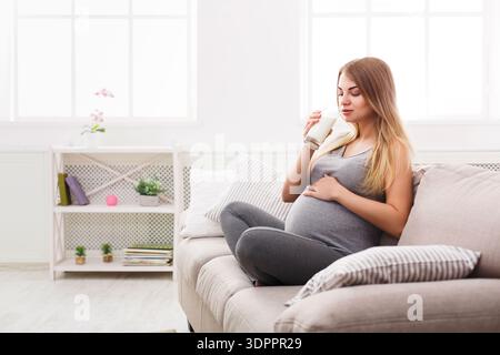 Schwangere Frau genießt einen Drink, während sie auf einer Couch in einem hellen Raum sitzt Stockfoto