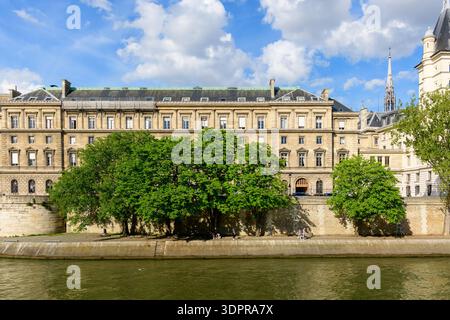 Paris, Frankreich - 14. April 2022: Das Äußere von Le Tribunal Judiciaire ist hinter Laubbäumen entlang der seine zu sehen, wo Menschen unter teilweise bewölktem Himmel auf der Uferpromenade sitzen und gehen. Stockfoto