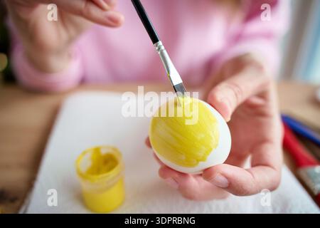Nahaufnahme von Frauenhänden, die weißes Ei halten, während sie es mit gelber Farbe und Pinsel auf dem Tisch streichen. Ostervorbereitung und Eierdekoration für die Feiertage Co Stockfoto