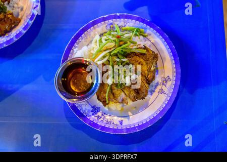 Ho-Chi-Minh-Stadt, Vietnam - 13. Januar 2020: Blick von oben auf einen Teller vietnamesisches gegrilltes Schweinekotelett serviert auf Reis mit eingelegtem Gemüse und einer kleinen Schüssel Dippsoße auf einem blauen Tisch. Stockfoto