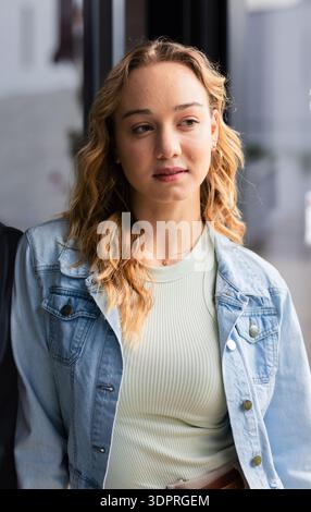 Frau, die an der Glasplatte in der Lobby stand und nach rechts blickte, während sie eine Jeansjacke und ein geripptes Oberteil trug Stockfoto