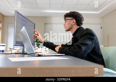 Asiatischer Mann mit Brille, der Daten auf dem Monitor analysiert, Laptop neben Kaffeetasse im Büro, Kopierraum Stockfoto
