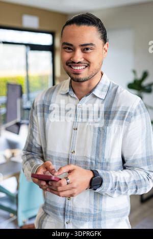Mittelgroßer asiatischer Mann lächelt, während er ein rotes Smartphone hält und Smartwatch in einem modernen Büro trägt Stockfoto