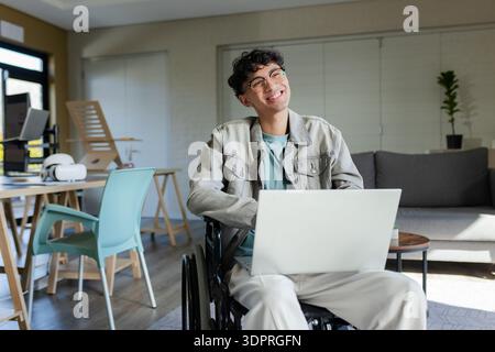 Lächelnder Mann, der im Rollstuhl sitzt und an einem Laptop in einem modernen Wohnzimmer mit VR-Headset arbeitet Stockfoto