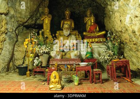 Mont Phousi, Laos - 24. März 2020: Mehrere Buddha- und Mönchstatuen sind in einem Höhlenschrein mit Opfergaben, Blumen, rituellen Gegenständen und kleinen Tischen auf einem gemusterten Boden vor einem felsigen Hintergrund angeordnet. Stockfoto