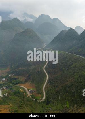 Eine schräge Aussicht aus der Luft zeigt eine gewundene Bergstraße, die steile bewaldete Karsthügel und ein schmales Tal im Norden Vietnams durchquert. Stockfoto